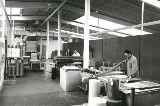 Black and white photo of a production hall in which two men are working at different workstations.